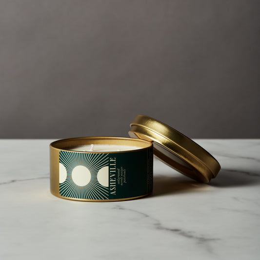 Asheville candle in a gold tin with a green label on a marble surface