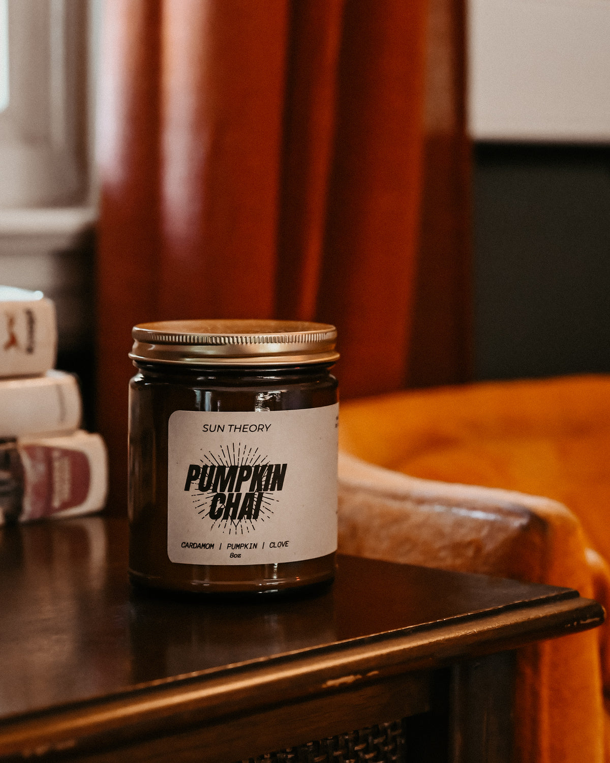 A jar candle labeled 'Pumpkin Chai' by Sun Theory sits on a dark wooden table in a cozy indoor setting with warm orange curtains and a leather chair in the background.