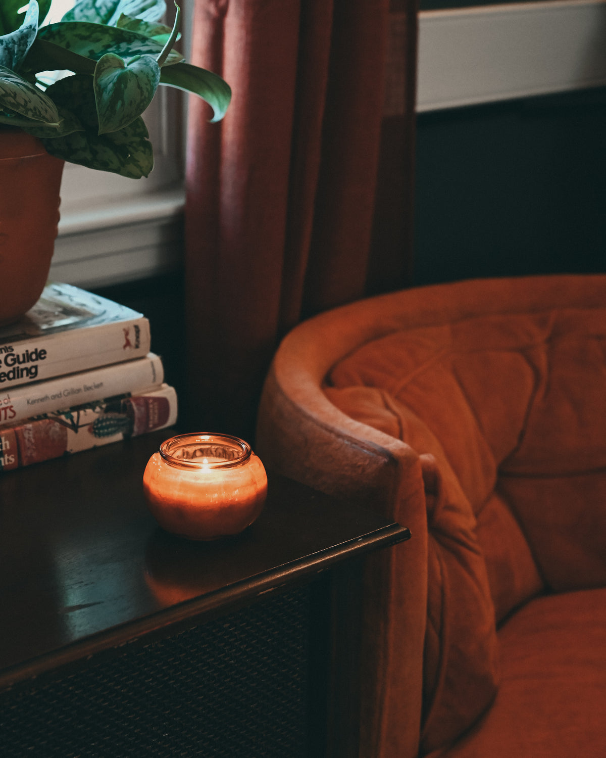 A lit orange candle in a small amber glass jar sits on a dark wooden side table next to a stack of books and a potted plant, with a rust-colored armchair and curtains in the background.