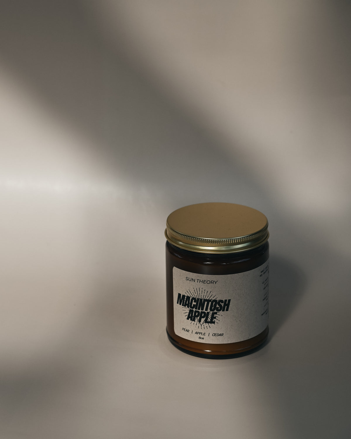 A single amber glass jar candle with a gold metal lid and a white label reading 'Sun Theory' and 'Macintosh Apple', photographed on a light gray surface with dramatic diagonal shadows.