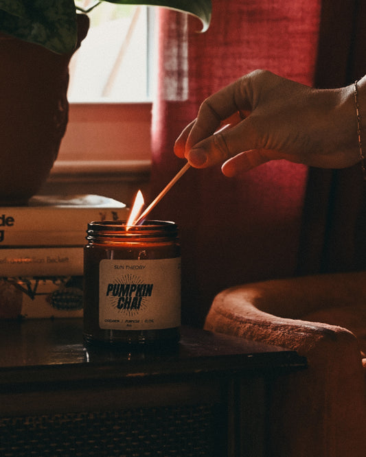 A hand lighting a candle with a wooden match in a cozy indoor setting. The candle is in an amber glass jar with a white label that reads 'Pumpkin Chai'
