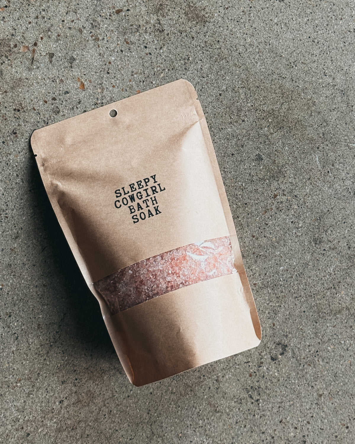 A kraft paper stand-up pouch labeled 'SLEEPY COWGIRL BATH SOAK' with a clear window showing pink bath salts, placed on a textured gray concrete surface.