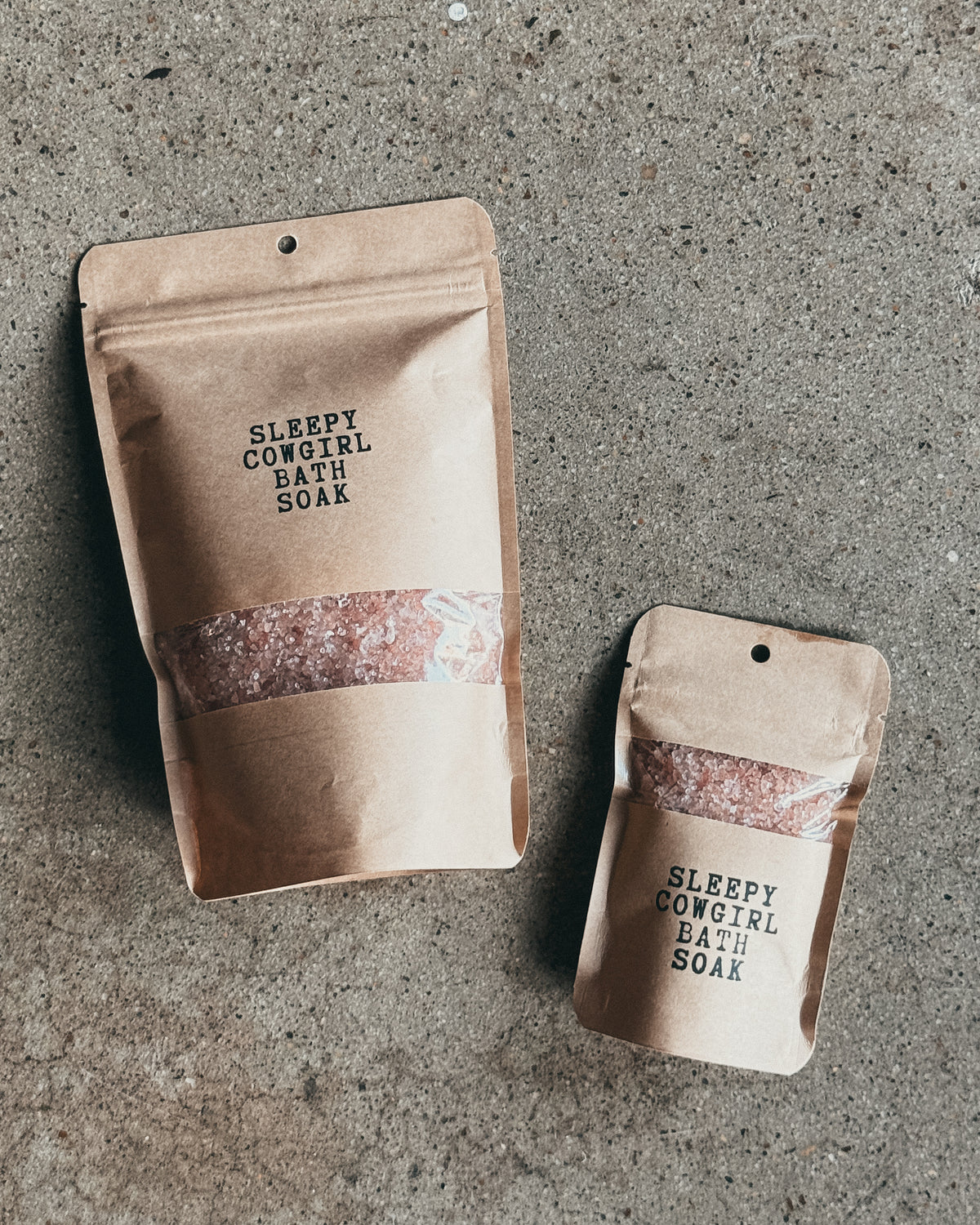 Two kraft paper stand-up pouches labeled 'SLEEPY COWGIRL BATH SOAK' with a clear window showing pink bath salts, placed on a textured gray concrete surface.