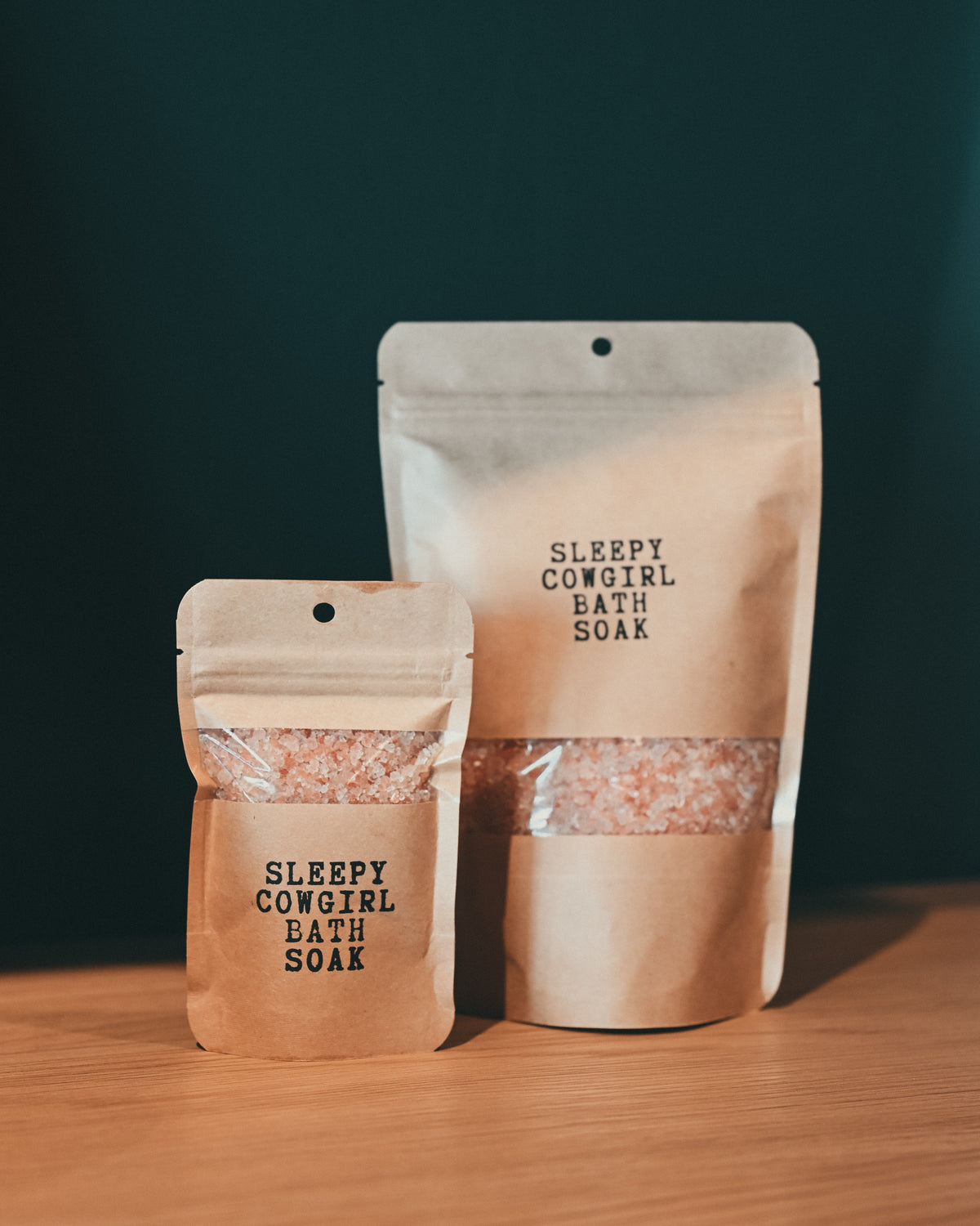 Two kraft paper pouches of Sleepy Cowgirl Bath Soak, one small and one large, standing on a wooden surface against a dark green background.