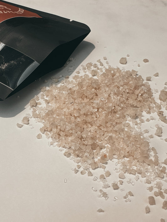 Heap of pink salt crystals next to a black bath soak packaging on a light surface