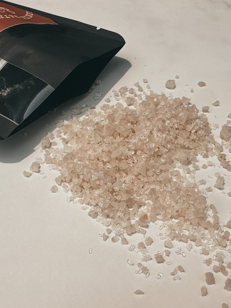 Heap of pink salt crystals next to a black bath soak packaging on a light surface