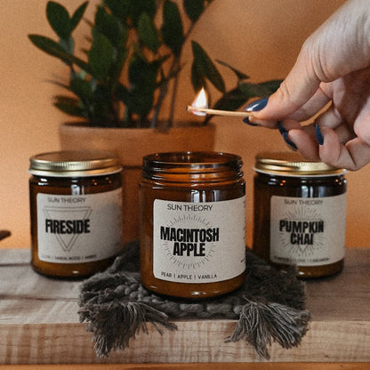 Trio of fall candle jars being lit by a hand focused on a Macintosh Apple candle