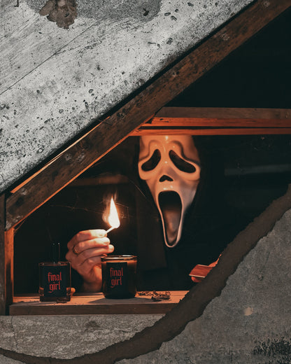 A person wearing a Ghostface-style mask sits in a dark attic window, holding a lit match near small bottles. The scene is framed by rough concrete walls and wooden framing, with dramatic low lighting and a horror aesthetic.