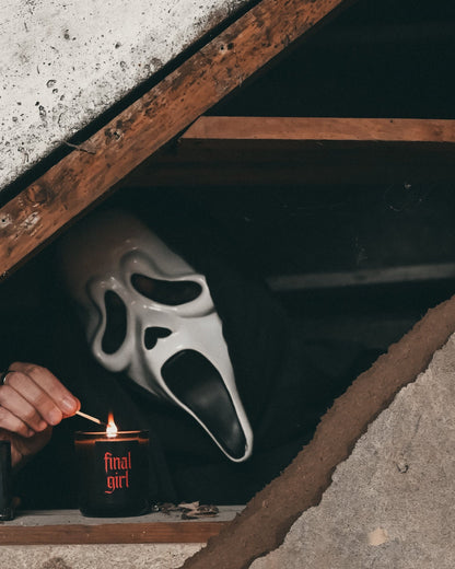 A person wearing a Ghostface-style mask sits in a dark basement space beneath a concrete roof, lighting a candle with a match. A black candle with red text reading 'Final Girl' are visible on the shelf.