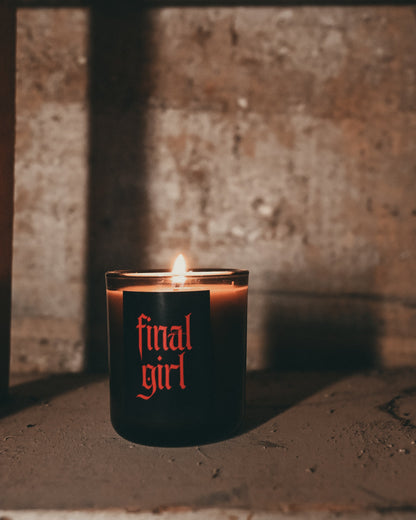 A black candle with a lit flame sits on a rustic wooden shelf. The candle features the words 'final girl' printed vertically in a red serif font.