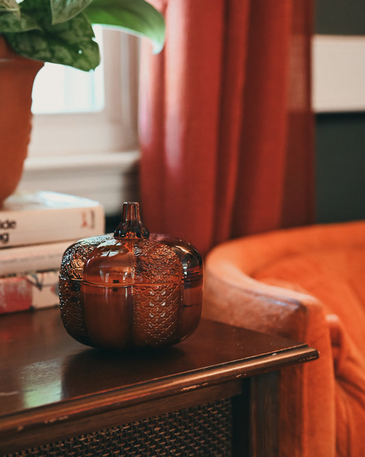 A textured amber glass pumpkin-shaped container with a lid, styled on a dark wooden side table in a cozy room setting with rust-orange curtains and a plant in the background.