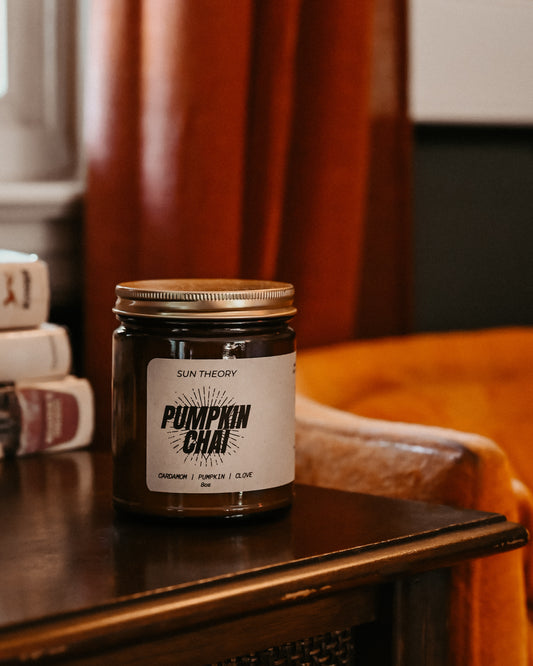 A jar candle labeled 'Pumpkin Chai' by Sun Theory sits on a dark wooden table in a cozy indoor setting with warm orange curtains and a leather chair in the background.