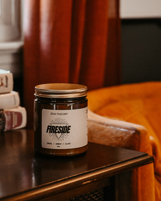 A glass jar candle with a gold metal lid and a white label reading 'FIRESIDE' and 'SUN THEORY' sits on a dark wooden table in a cozy indoor setting with warm orange and brown tones.