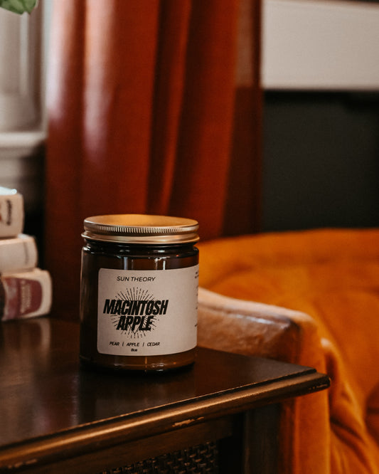 A jar candle labeled 'Sun Theory' and 'Macintosh Apple' sits on a dark wooden table in a cozy indoor setting with warm red curtains and an orange couch in the background.
