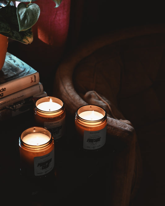 Three lit candles in amber glass jars styled on a dark wooden surface next to stacked books and a plant in a dimly lit room.