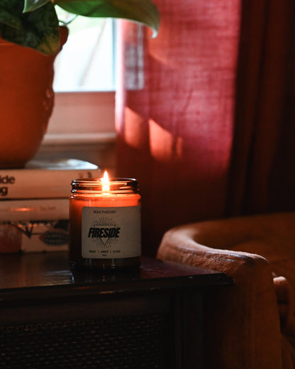 A lit candle in a dark glass jar sits on a wooden table in a cozy indoor setting with warm red curtains, a potted plant, and stacked books in the background. The label reads 'FIRESIDE' and 'Sun Theory".
