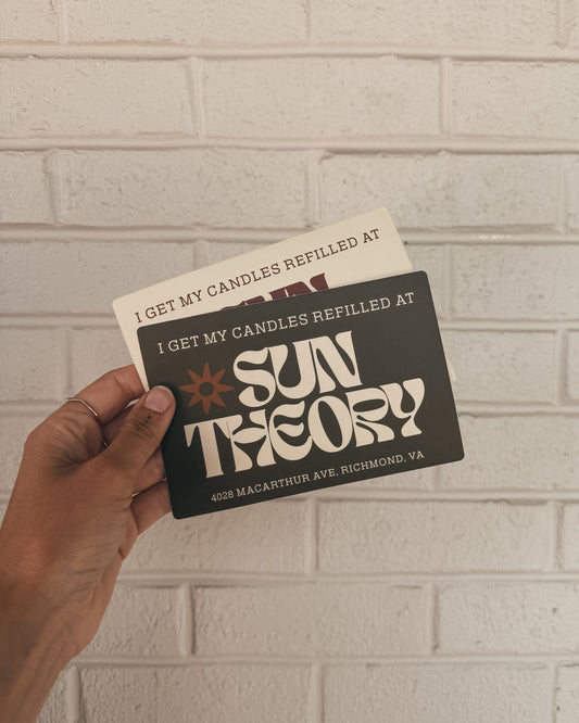 Two bumper stickers that read 'I get my candles refilled at Sun Theory' with the address "4028 Macarthur Ave, Richmond VA" at the bottom of the sticker held up against a light brick wall background.