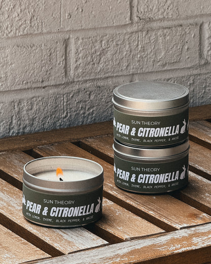 Three silver metal tins of Sun Theory Pear & Citronella candles arranged on a rustic wooden slatted surface against a light gray brick wall. One tin is open with a lit candle, and two tins are stacked.