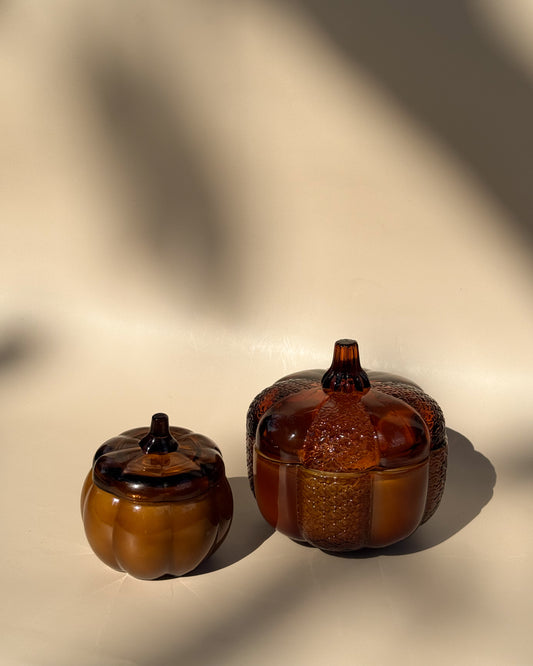 Two amber glass pumpkin-shaped containers with lids, one small and one larger, displayed on a warm beige surface with soft shadows.