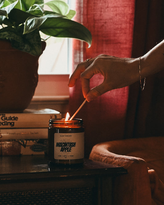 A lit candle in a dark glass jar sits on a wooden table in a warm, dimly lit room. A hand holds a match above the flame.