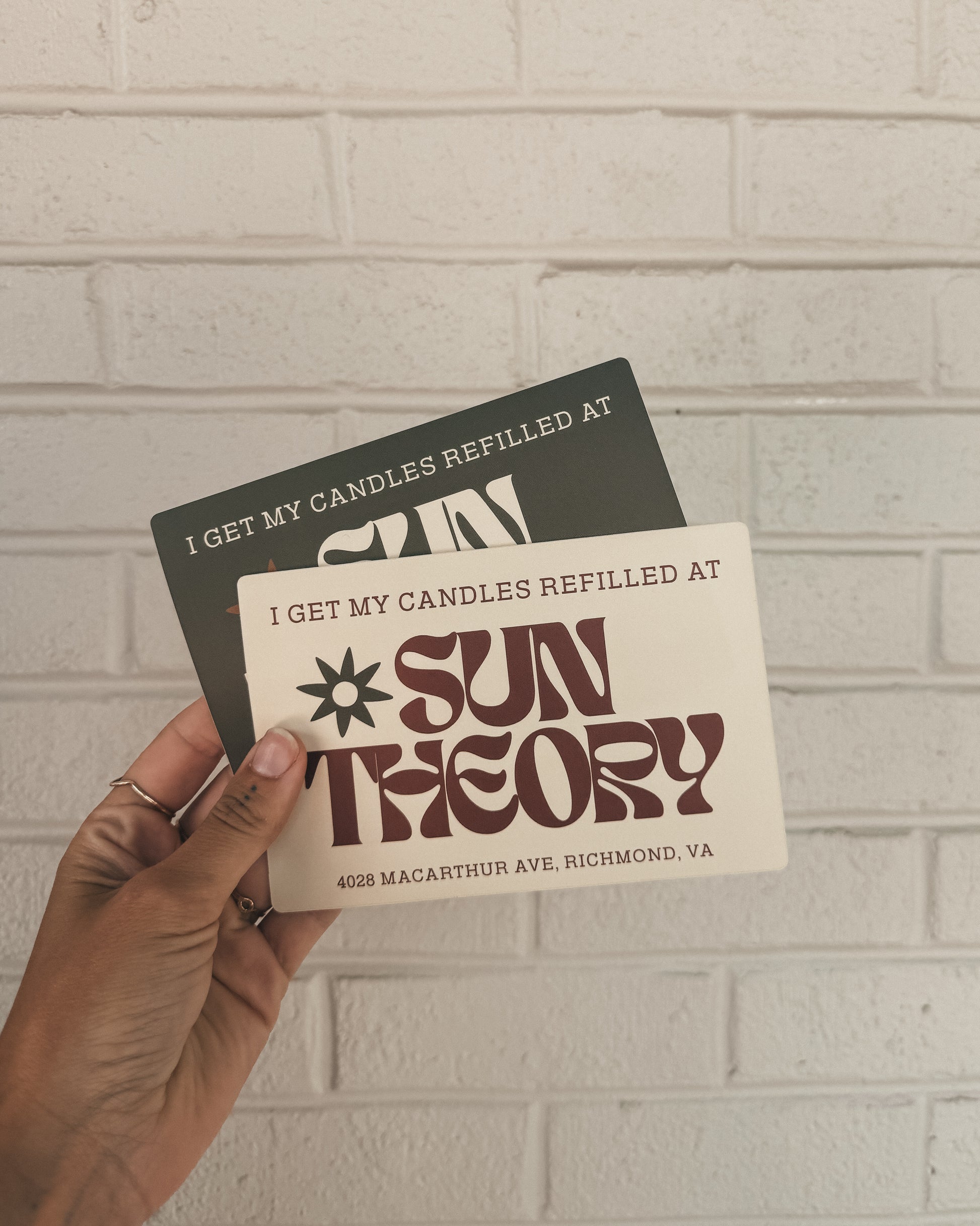 Two bumper stickers that read 'I get my candles refilled at Sun Theory' with the address "4028 Macarthur Ave, Richmond VA" at the bottom of the sticker held up against a light brick wall background.