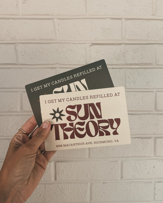 Two bumper stickers that read 'I get my candles refilled at Sun Theory' with the address "4028 Macarthur Ave, Richmond VA" at the bottom of the sticker held up against a light brick wall background.