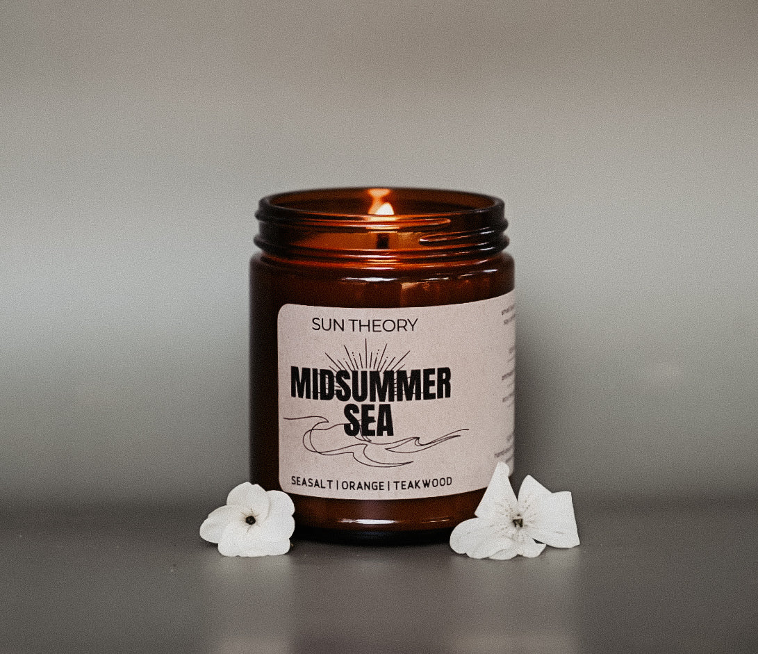 Candle in an amber jar labeled 'Midsummer Sea' by Sun Theory on a neutral background with white flowers.