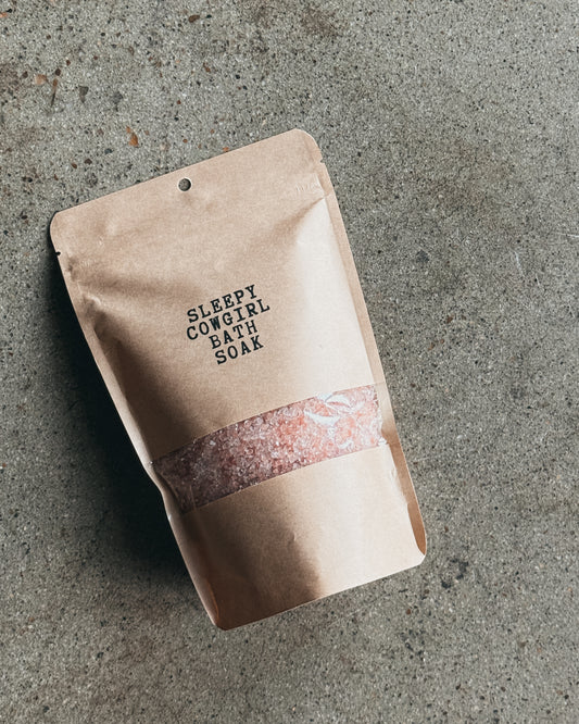 A kraft paper stand-up pouch labeled 'SLEEPY COWGIRL BATH SOAK' with a clear window showing pink bath salts, placed on a textured gray concrete surface.