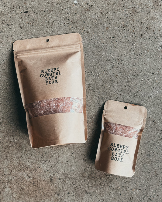 Two kraft paper stand-up pouches labeled 'SLEEPY COWGIRL BATH SOAK' with a clear window showing pink bath salts, placed on a textured gray concrete surface.
