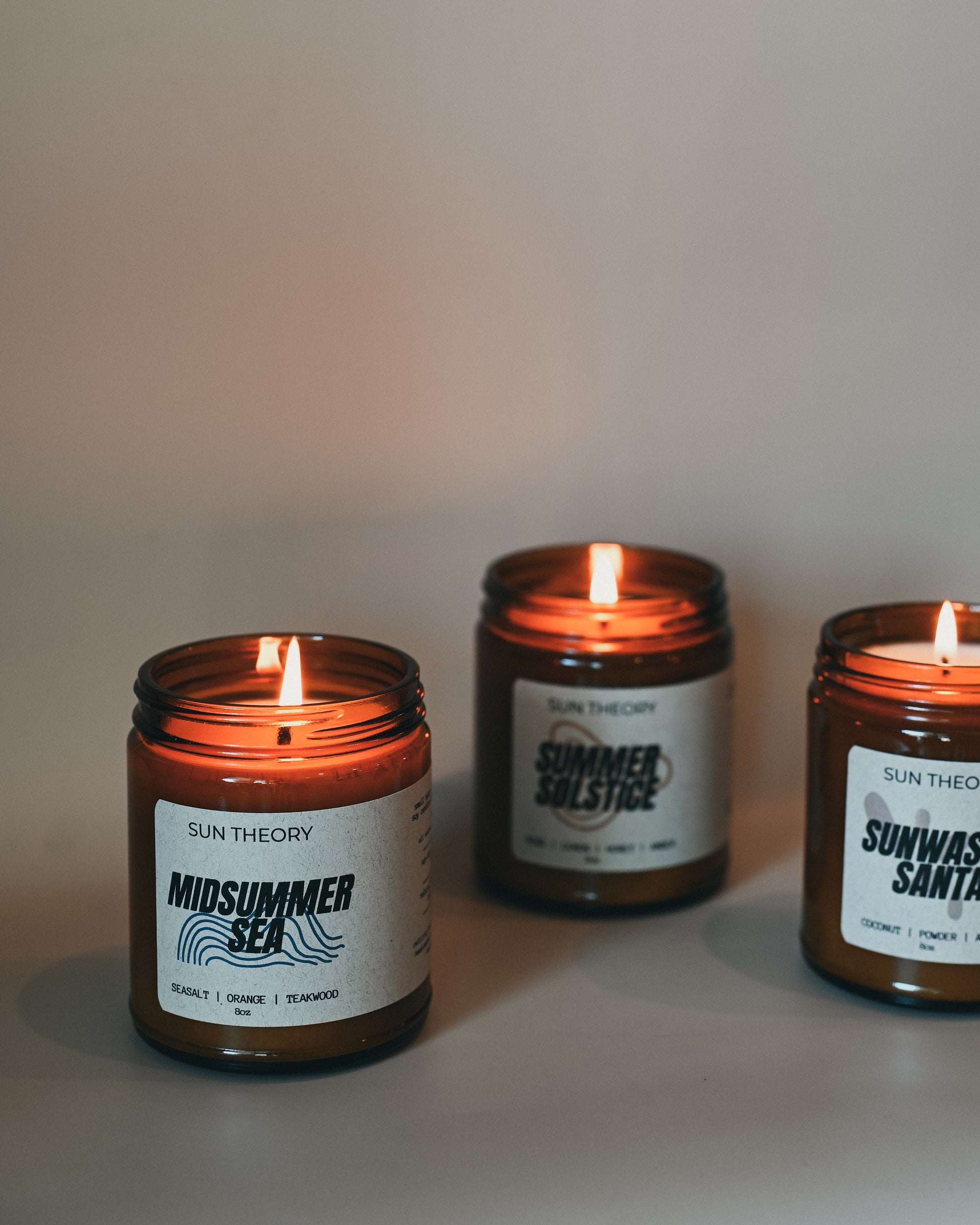 Three lit candles in brown jars with labels on a neutral background