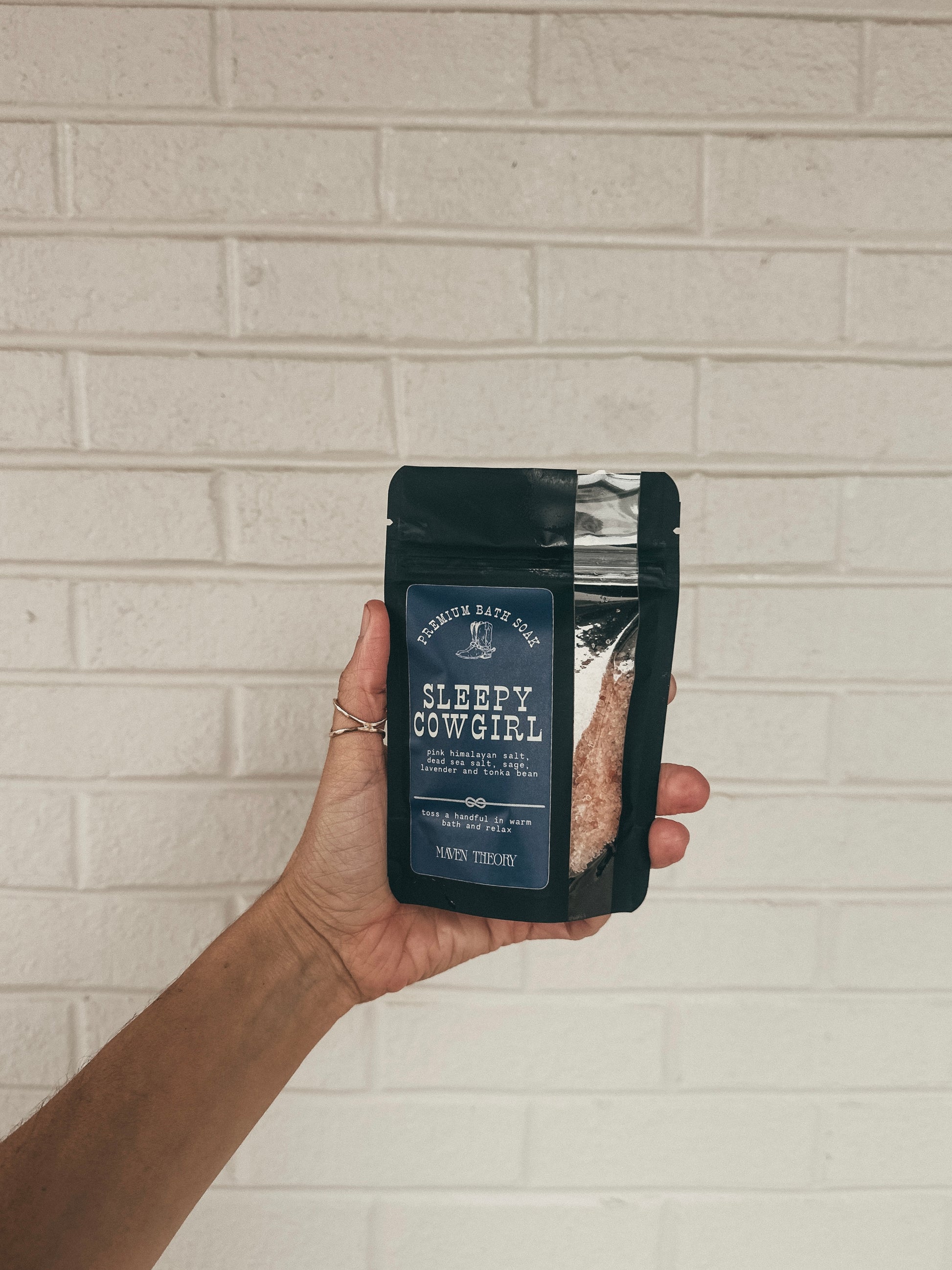 Hand holding a package labeled 'Sleepy Cowgirl' against a light brick wall.
