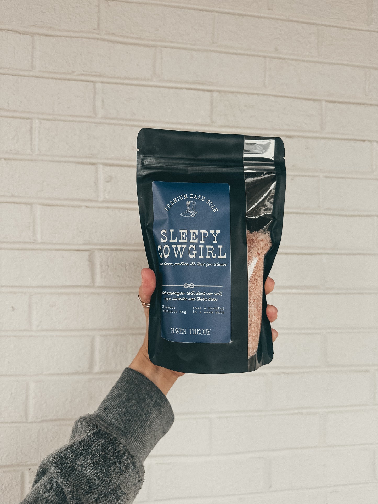 Hand holding a package of 'Sleepy Cowgirl' bath soak against a light brick wall.