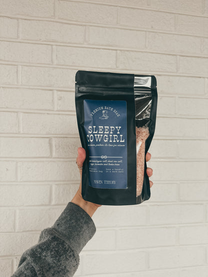 Hand holding a package of 'Sleepy Cowgirl' bath soak against a light brick wall.
